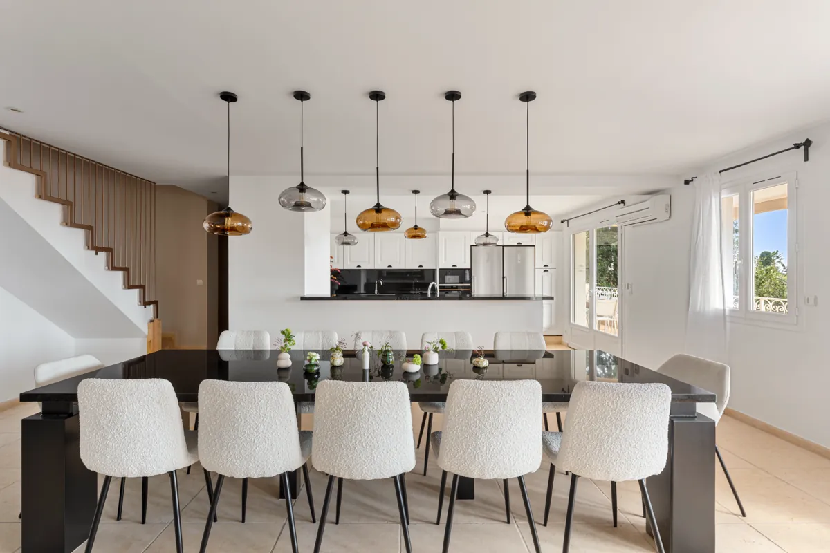 Salle à manger avec une longue table noire et des chaises blanches, suspensions en verre ambré, cuisine ouverte au fond, escalier à gauche.