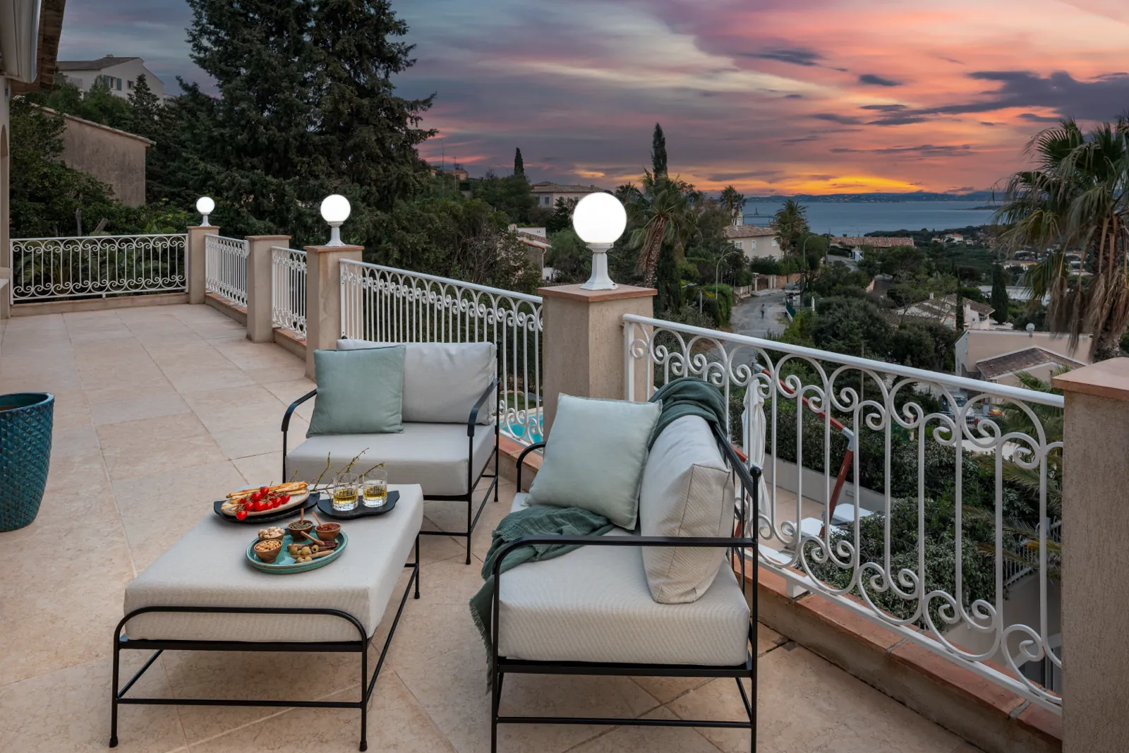 Terrasse dallée au couchant, deux fauteuils bas et une table basse avec des assiettes, balustrade blanche en fer forgé, mer au loin sous un ciel orangé.
