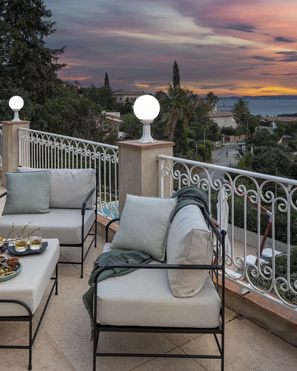 Terrasse dallée au couchant, deux fauteuils bas et une table basse avec des assiettes, balustrade blanche en fer forgé, mer au loin sous un ciel orangé.