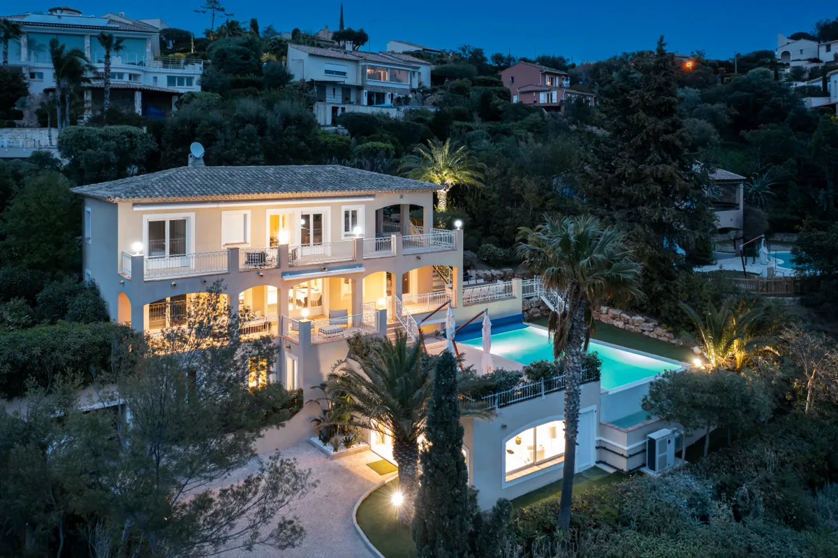 Vue aérienne de la villa à la tombée de la nuit, façade éclairée, piscine allumée, palmiers et pins autour.