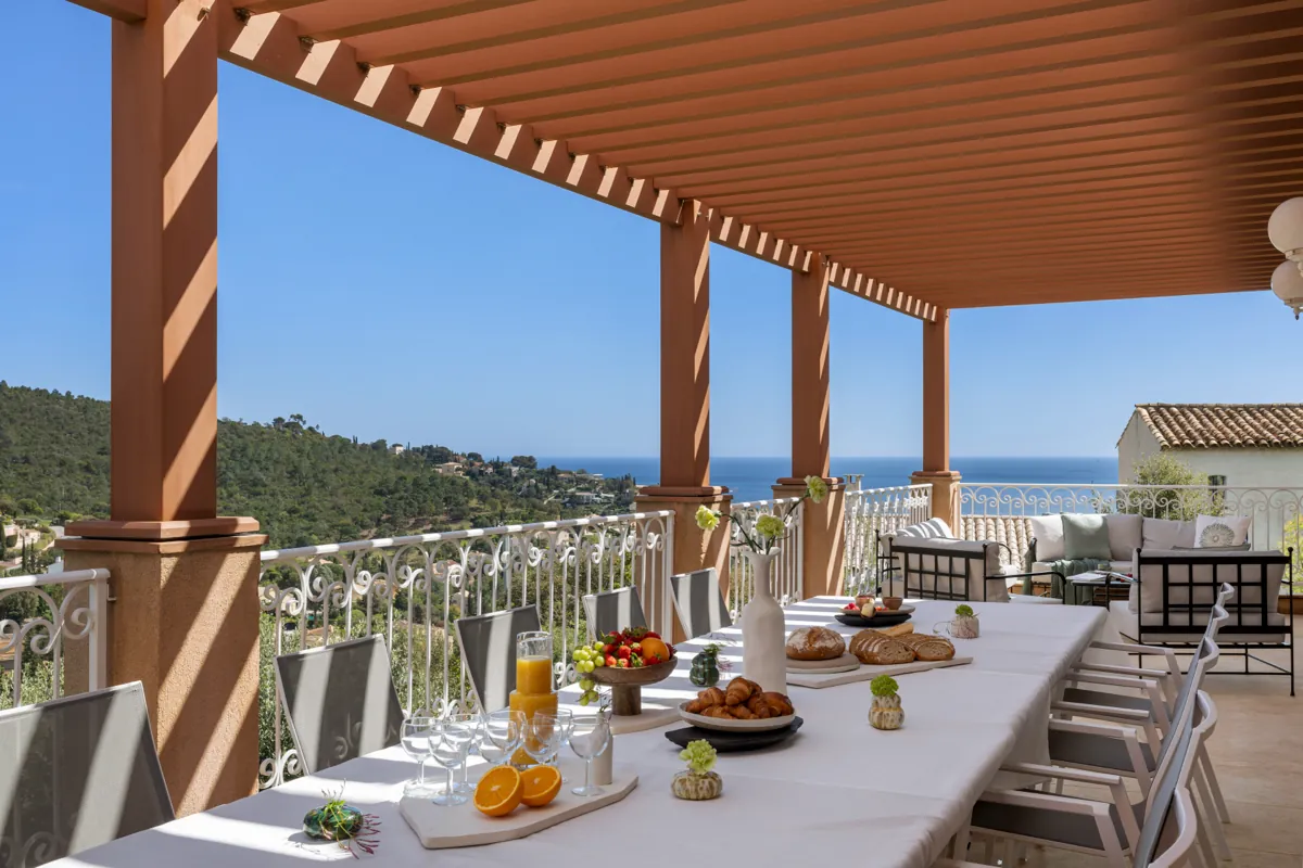 Terrasse couverte à colonnes ocre, table dressée pour un repas, collines boisées et mer au fond.