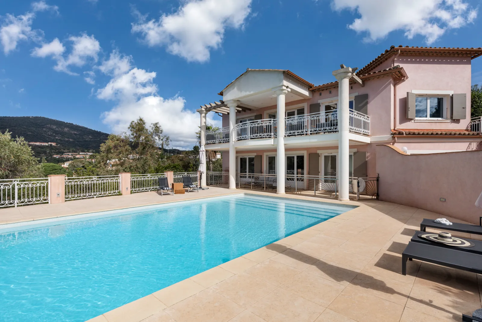 Villa rose pâle à colonnes blanches, piscine au premier plan, collines derrière.